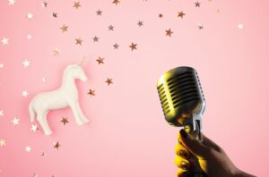 Hand holding a vintage microphone with unicorn figurine and star confetti on a pastel pink background illustrating creativity in podcast sponsorship.