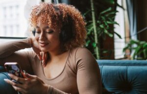Young woman with curly hair wearing headphones and smiling while listening to a podcast on her smartphone.