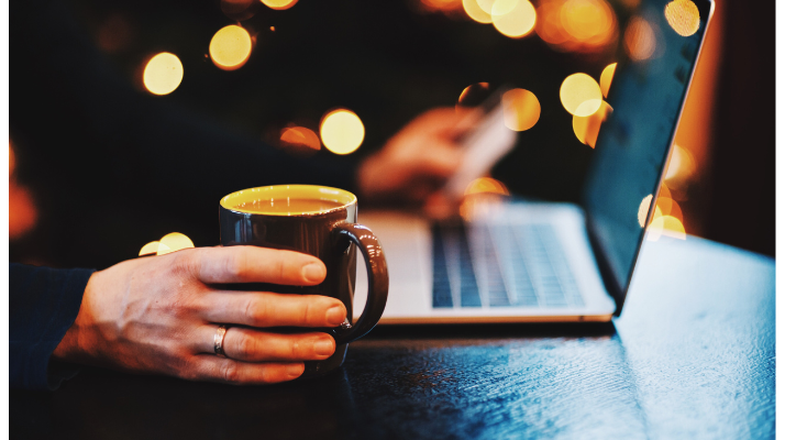 An image showing a laptop on a table, male hand holding a cup of coffee with a dark, but festive background. Podcast Advertising 2024