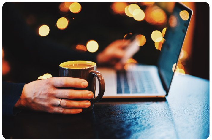 An image showing a laptop on a table, male hand holding a cup of coffee with a dark, but festive background. Podcast Advertising 2024