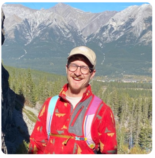 Man smiling outdoors with mountains and trees in the background.