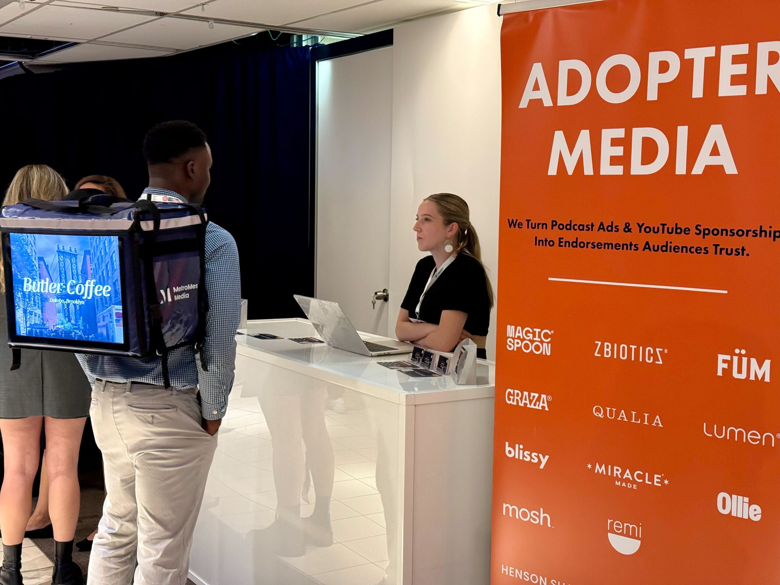 ADOPTER Media exhibit booth at Advertising Week New York 2025 with branded signage and event attendees discussing podcast and YouTube advertising.