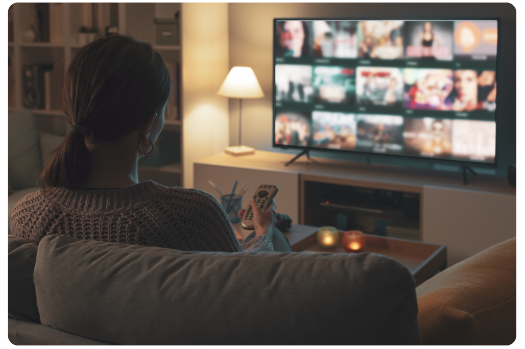 A woman sitting on a sofa in a cozy living room, holding a remote control and watching a smart TV displaying a streaming platform interface.