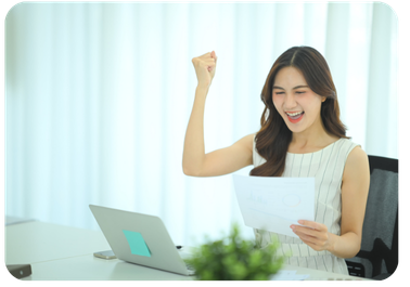 Woman at a desk celebrating with a raised fist while holding a printed report, with a laptop in front of her