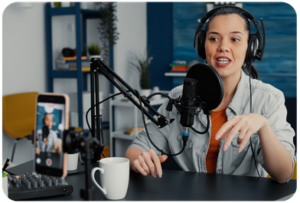 Female podcast host recording at a microphone with headphones on, with a smartphone camera in view capturing the session
