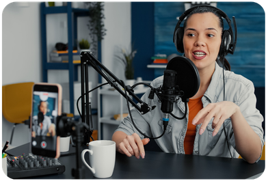Female podcast host recording at a microphone with headphones on, with a smartphone camera in view capturing the session