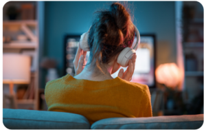 Woman at home on her couch wearing headphones while watching a podcast on her TV, illustrating the rise in at-home podcast listening in 2026.