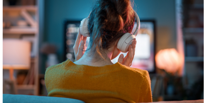 Woman at home on her couch wearing headphones while watching a podcast on her TV, illustrating the rise in at-home podcast listening in 2026.