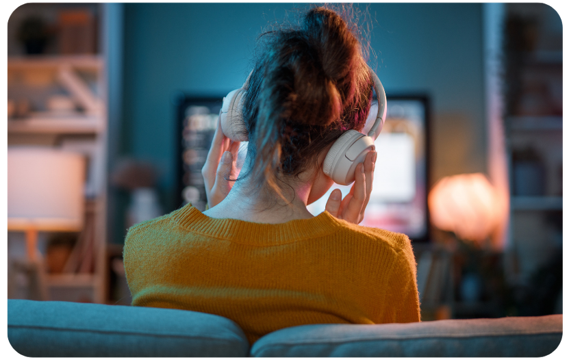Woman at home on her couch wearing headphones while watching a podcast on her TV, illustrating the rise in at-home podcast listening in 2026.