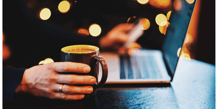 An image showing a laptop on a table, male hand holding a cup of coffee with a dark, but festive background. Podcast Advertising 2024