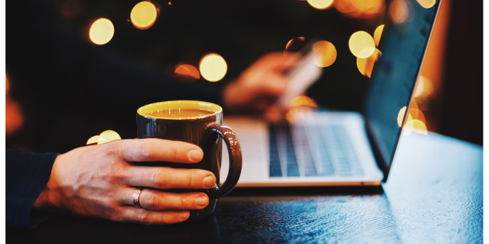 An image showing a laptop on a table, male hand holding a cup of coffee with a dark, but festive background. Podcast Advertising 2024