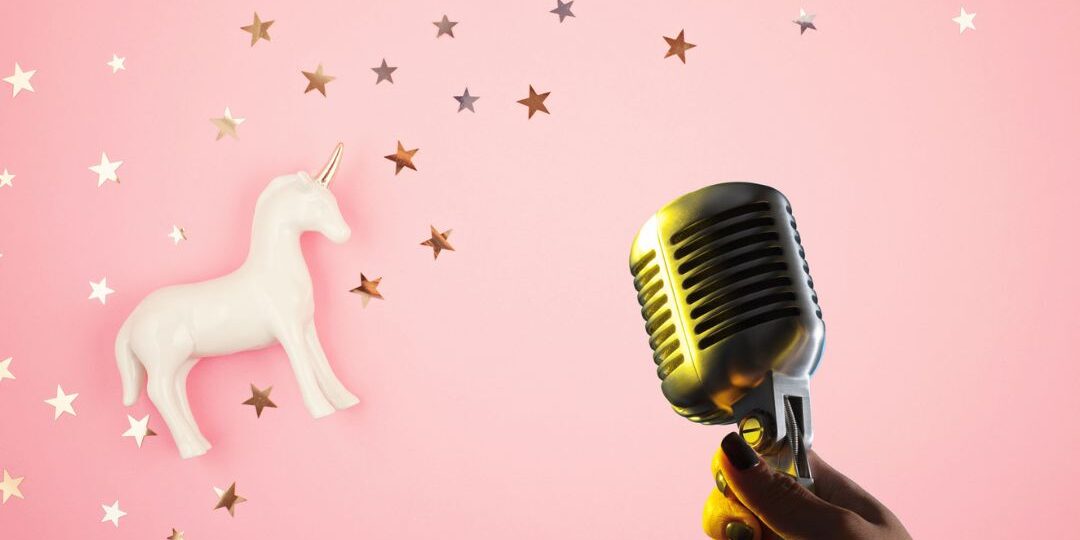 Hand holding a vintage microphone with unicorn figurine and star confetti on a pastel pink background illustrating creativity in podcast sponsorship.