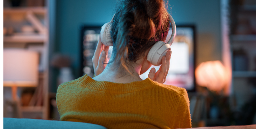 Woman at home on her couch wearing headphones while watching a podcast on her TV, illustrating the rise in at-home podcast listening in 2026.