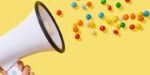 Megaphone dispersing colorful confetti and sprinkles on bright yellow background representing message amplification and promotional impact
