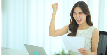 Woman at a desk celebrating with a raised fist while holding a printed report, with a laptop in front of her