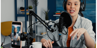 Female podcast host recording at a microphone with headphones on, with a smartphone camera in view capturing the session