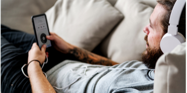 Man wearing headphones relaxing on a couch while listening to a podcast on his phone