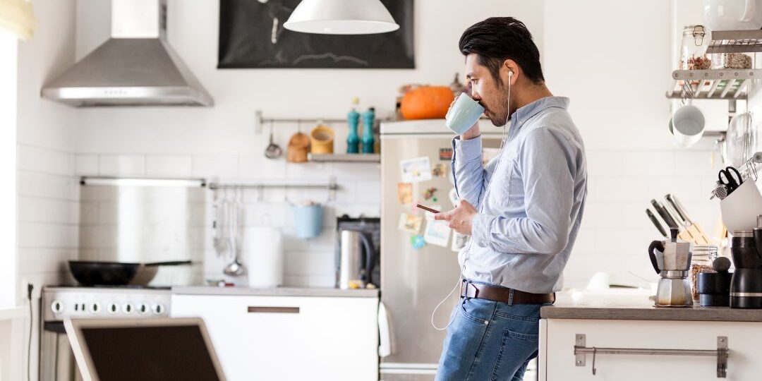 Man drinking coffee while checking phone in kitchen, representing modern digital lifestyle and direct response marketing engagement.