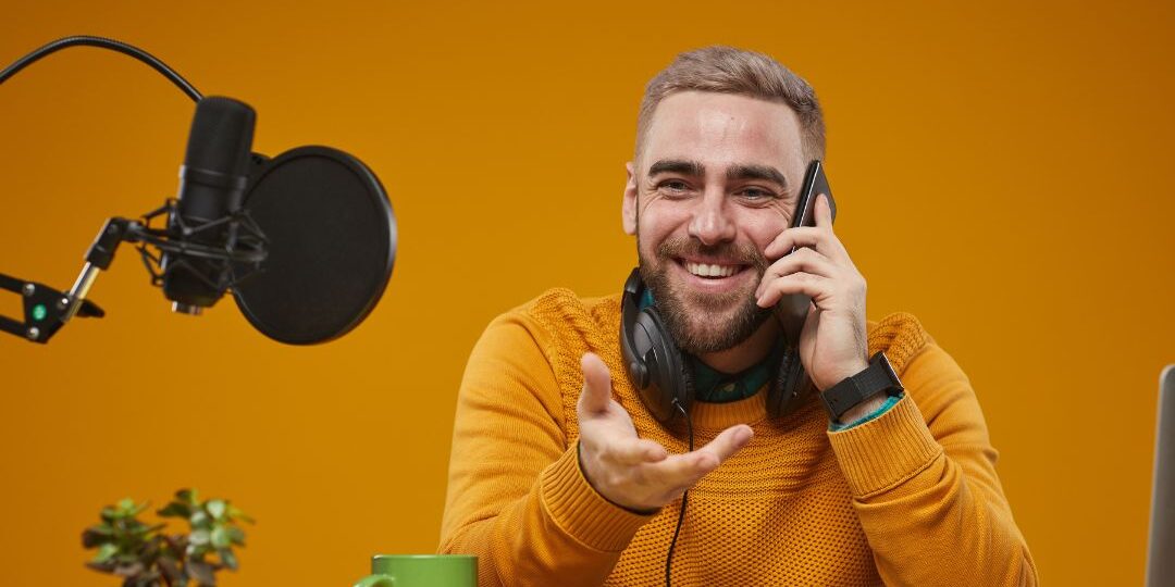Male podcaster wearing yellow sweater speaking on phone while recording with professional studio microphone and headphones on golden background